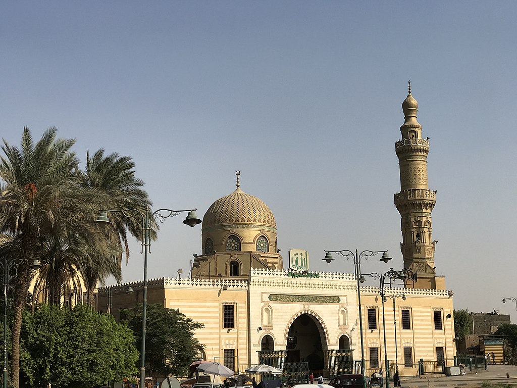 A mosque in urban Cairo with a golden dome and minaret, flanked to the left by palm trees and shrubs.